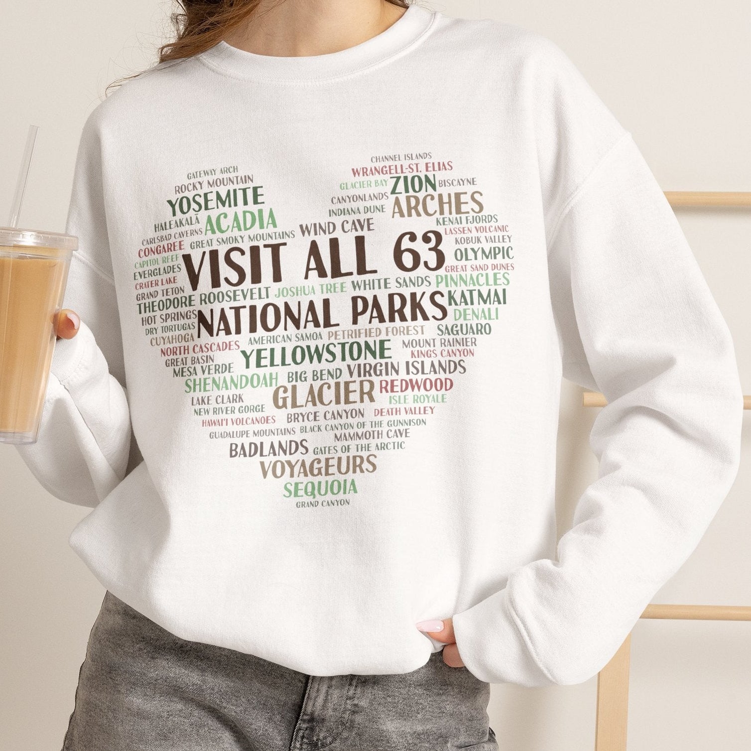 A woman wearing a white crewneck sweatshirt with a 'Visit All 63 National Parks' word heart graphic, paired with gray denim shorts and holding an iced coffee.