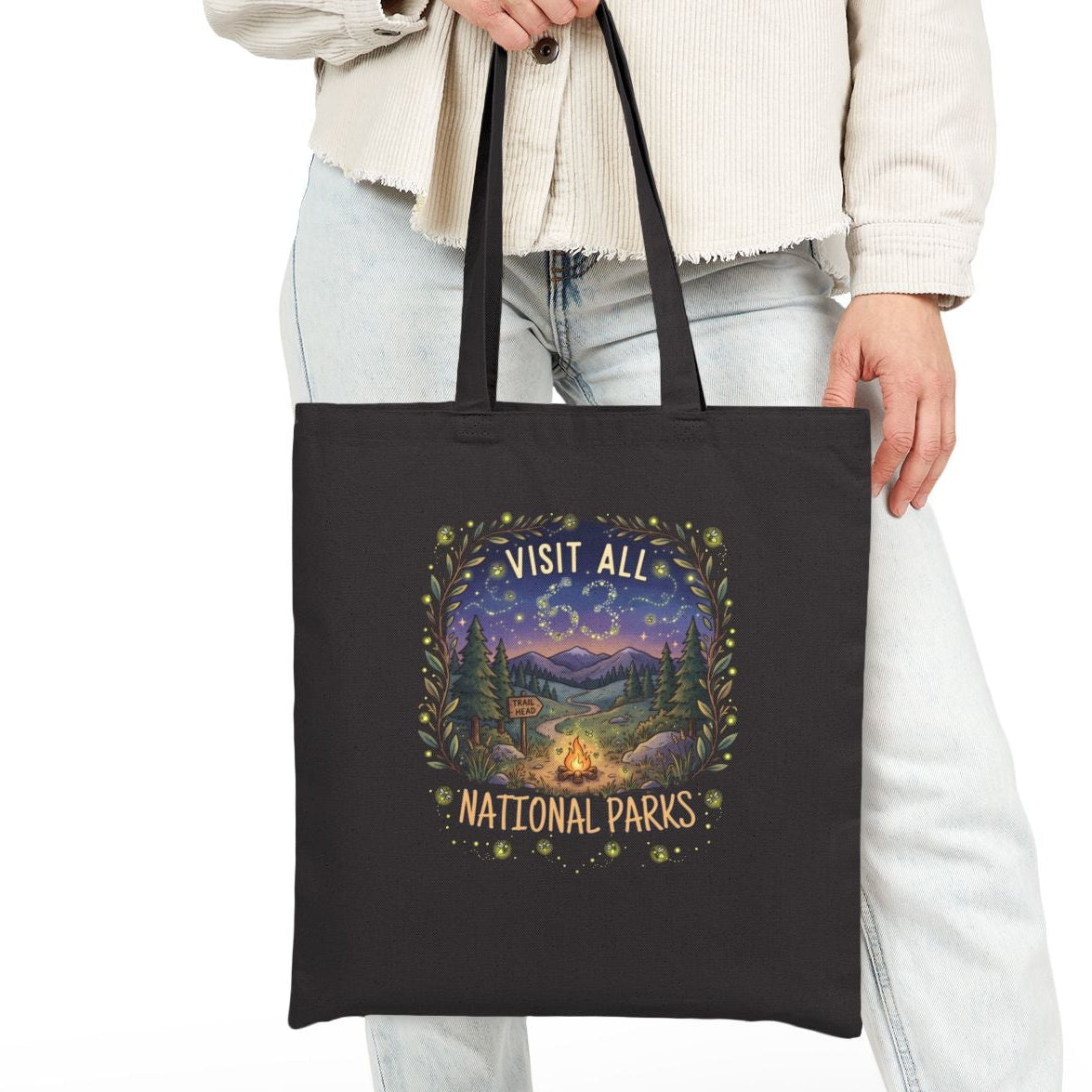 A person wearing a light beige jacket, light wash jeans, and white sneakers holds a black canvas tote bag with a colorful 'Visit All National Parks' graphic print on the front, set against a plain white studio background.