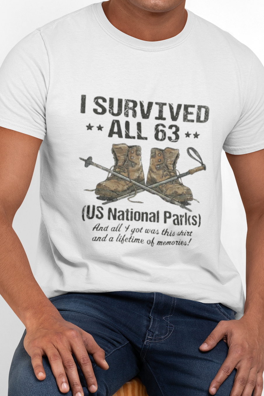 A man wearing a white graphic t-shirt that reads 'I SURVIVED ALL 63 (US National Parks) And all I got was laughter and a lifetime of memories!' with an illustration of hiking boots and a shovel, paired with dark blue jeans, seated on a wooden stool against a light gray background.