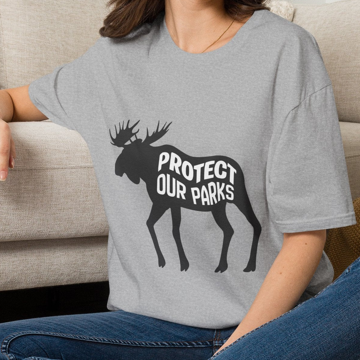 A woman sitting cross-legged on a woven rug in front of a beige sofa, wearing a heather gray t-shirt with a black moose graphic and the text 'PROTECT OUR PARKS'.