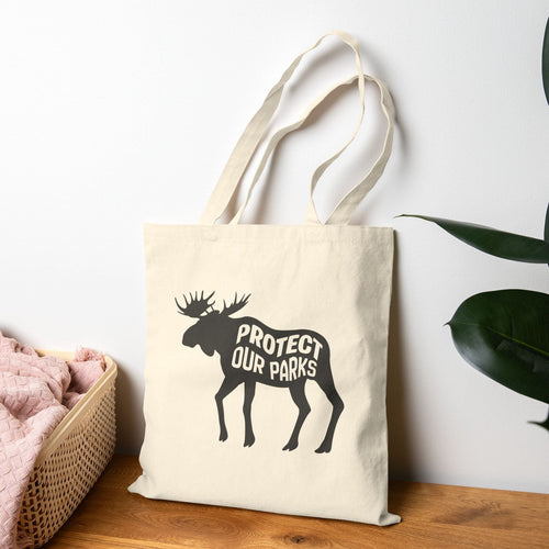 An off-white canvas tote bag with a black moose silhouette and the words 'PROTECT OUR PARKS' printed on the front, leaning on a wooden surface against a white wall with a pink blanket and green leaves nearby.