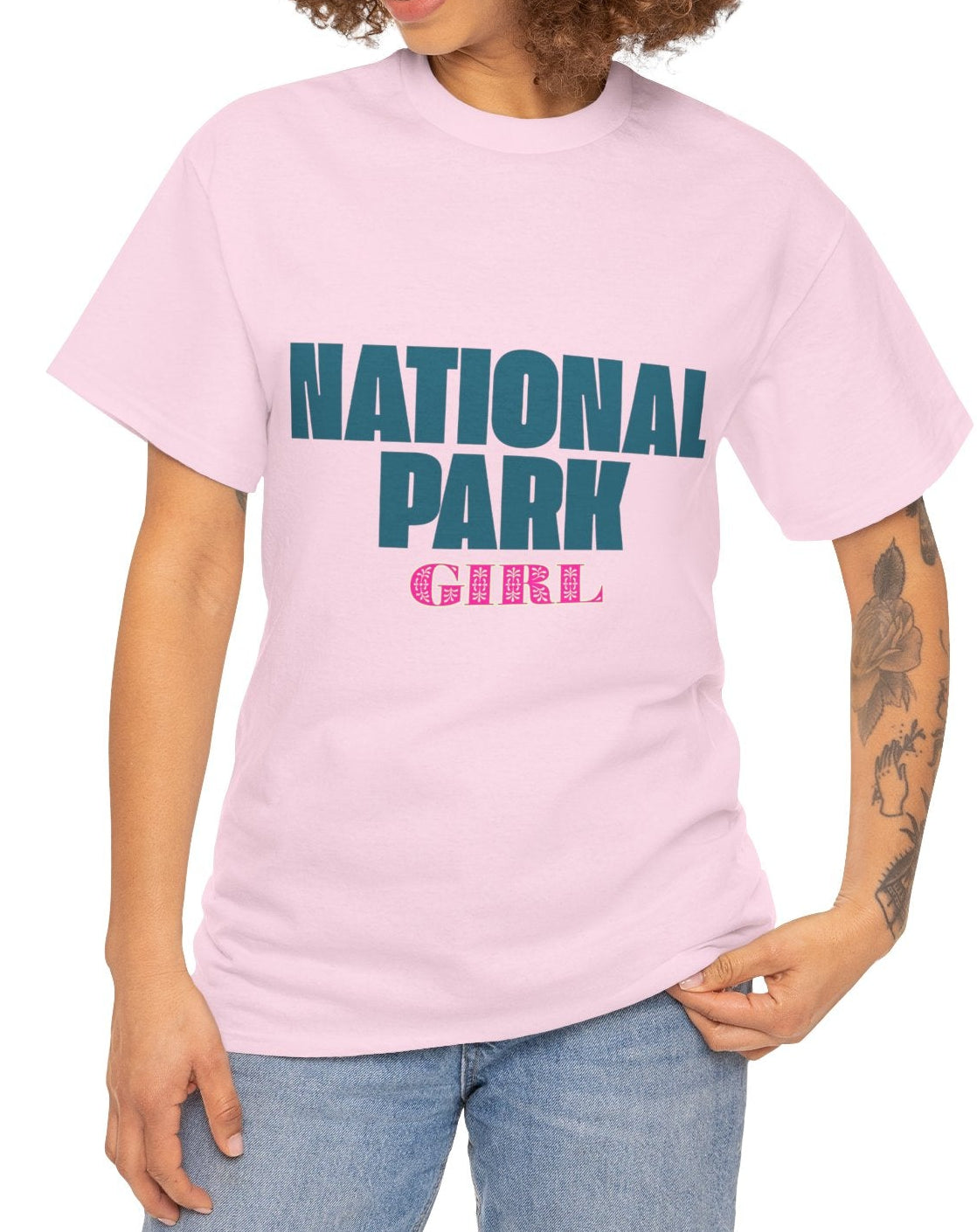 A woman with curly brown hair wearing a light pink t-shirt with 'NATIONAL PARK GIRL' printed on the front in teal and pink, paired with blue jeans, against a plain white background.