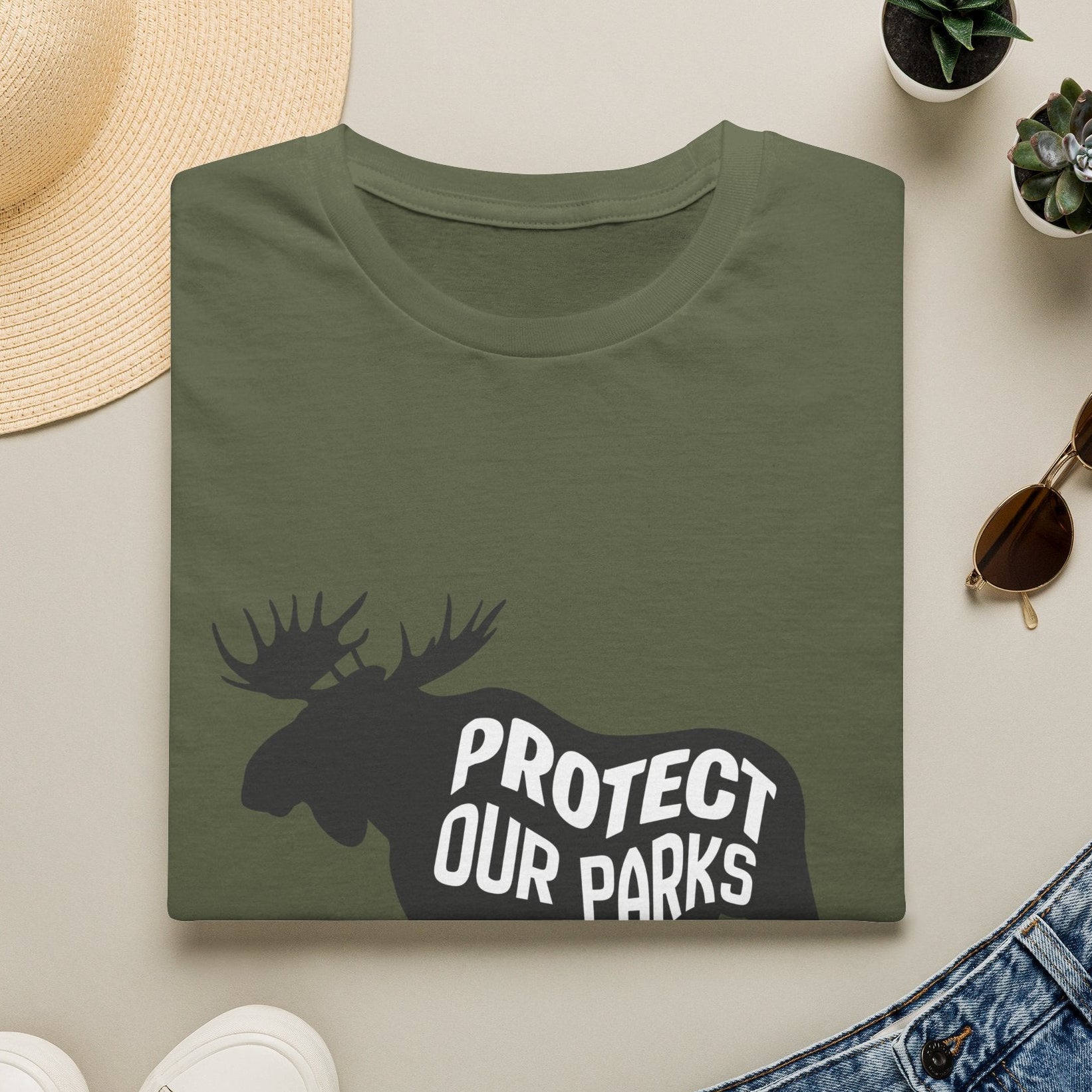 An olive green folded T-shirt with a black moose silhouette and the words 'PROTECT OUR PARKS' in white, styled in a flat lay with a straw hat, white sneakers, blue jeans, sunglasses, and small potted succulents on a light beige background.