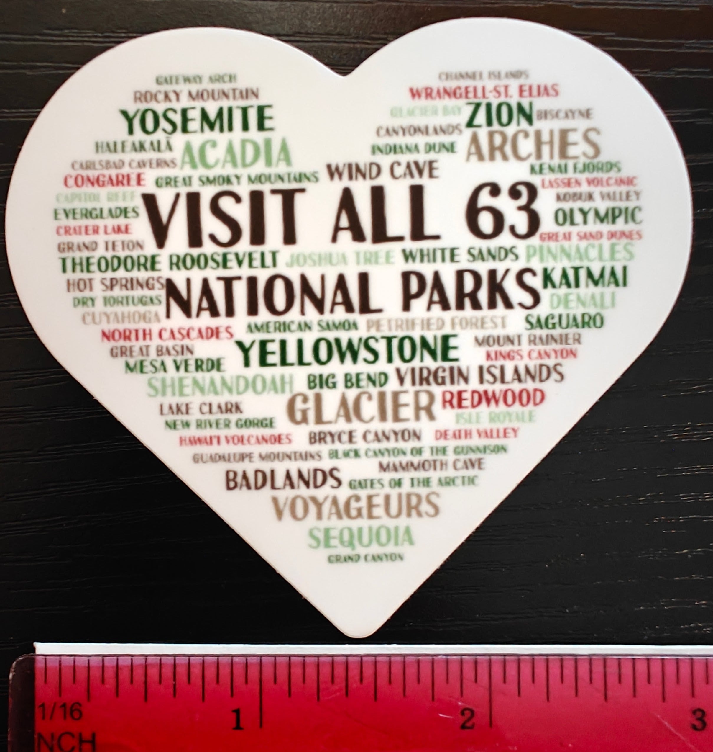A heart-shaped white sticker or sign featuring a 'Visit All 63 National Parks' word heart with various national park names in green and brown text, shown on a dark wood background with a red ruler at the bottom for scale.