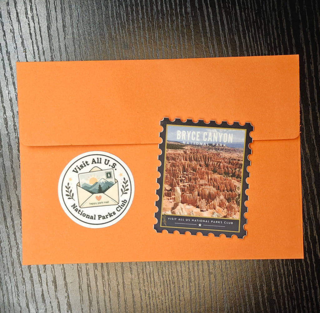 An orange envelope on a dark wood surface, featuring a die-cut postage stamp of Bryce Canyon National Park and a round white sticker that reads 'Visit All U.S.